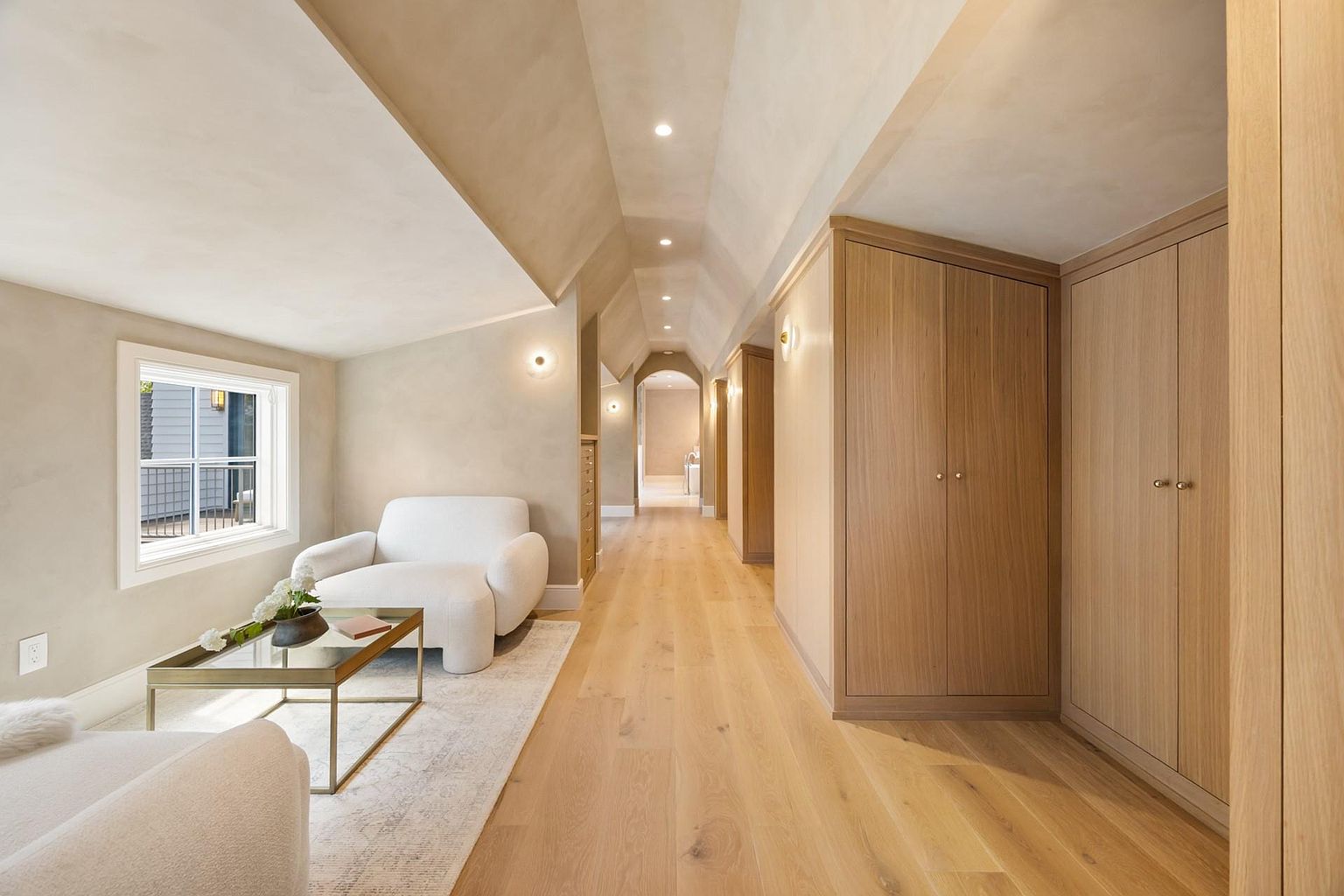 This interior shot showcases a long hallway with light wood flooring and a vaulted ceiling. Built-in wooden cabinets line one side of the hallway, while a seating area with a white armchair and glass-topped coffee table is visible on the left. The hallway leads to a distant archway, creating a sense of depth and spaciousness.