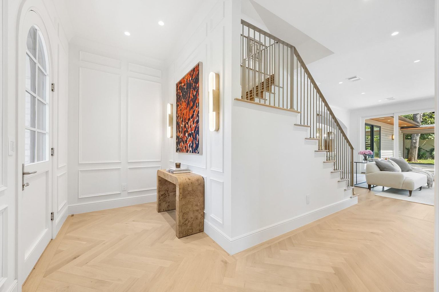 This is an interior shot of a bright and elegant entryway featuring a herringbone wood floor, white walls with decorative molding, and a staircase with a modern metal railing. An abstract painting hangs above a console table, adding a touch of color and sophistication to the space. The open layout leads into a living area with a view of the outdoors, creating a welcoming and airy atmosphere.