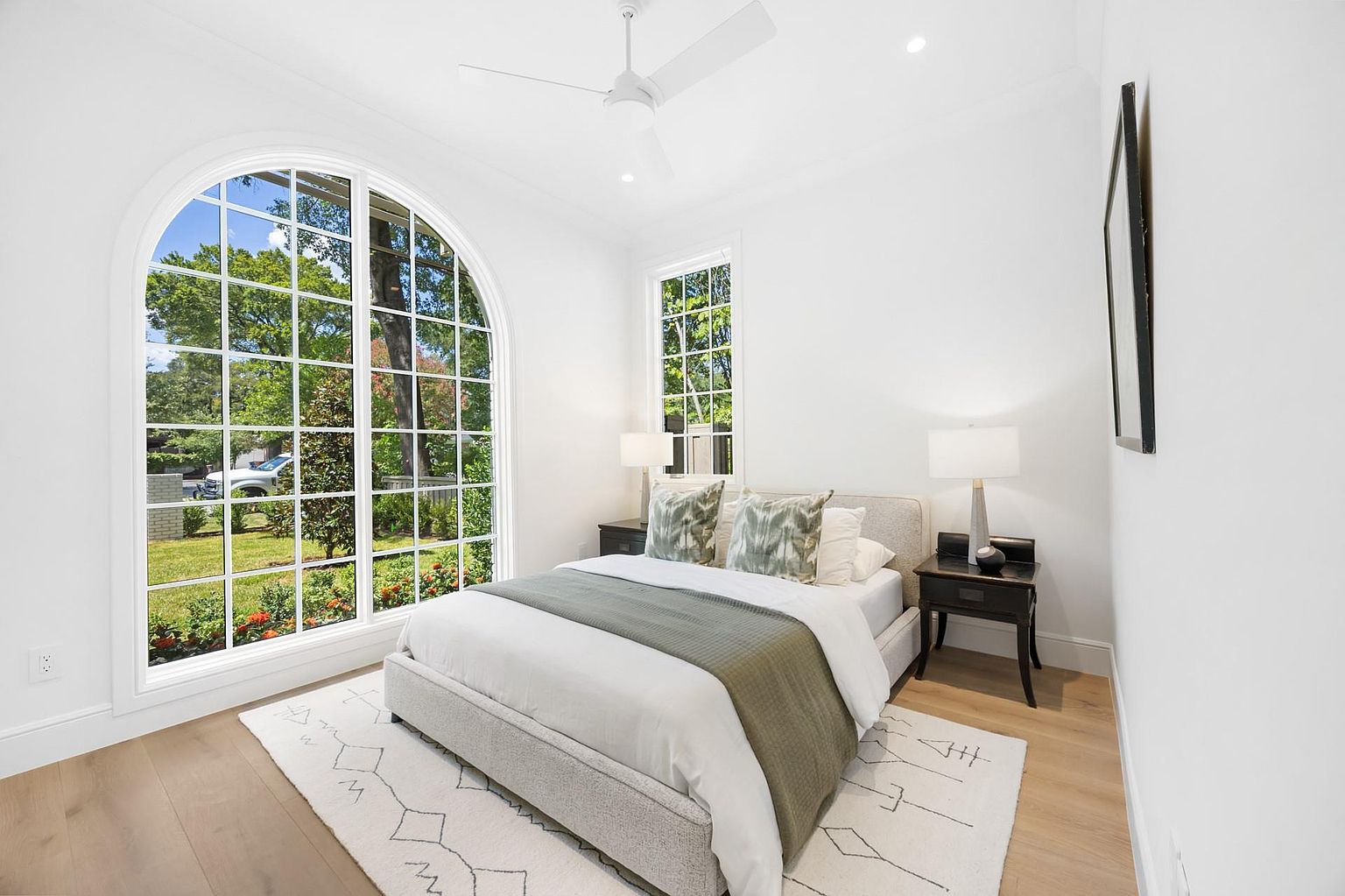 This is a bright and airy bedroom featuring a large arched window that provides ample natural light and a view of the outdoors. The room is decorated in a minimalist style with white walls, light wood flooring, and a neutral-toned bed. A geometric patterned rug adds a touch of visual interest to the space.