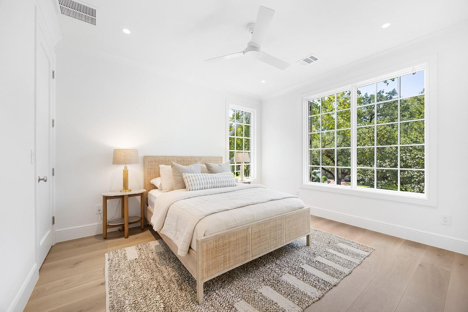 This is a bright and airy primary bedroom featuring a woven platform bed with neutral bedding, flanked by matching nightstands and lamps. Large windows provide ample natural light and views of the surrounding greenery. The room is decorated in a minimalist style with light wood flooring and a textured area rug, creating a serene and inviting atmosphere.