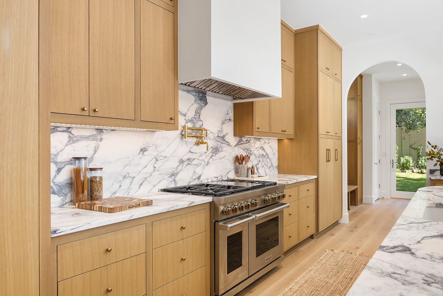This is a well-lit kitchen featuring light wood cabinetry with brass hardware, marble countertops and backsplash, and stainless steel appliances. A large range with a white hood is the focal point, and the open layout leads to an arched doorway. The overall impression is modern and luxurious.