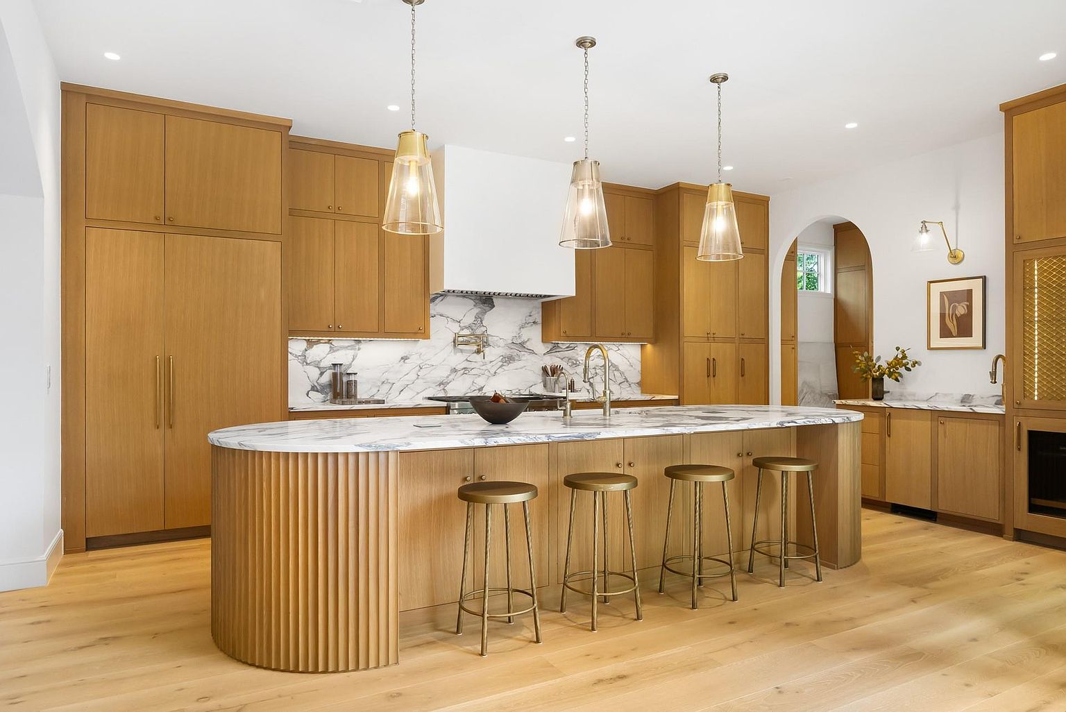 This is a bright and modern kitchen featuring custom wood cabinetry, marble countertops and backsplash, and a large island with seating. Three pendant lights hang above the island, providing ample lighting. The kitchen also features stainless steel appliances and a white range hood, creating a clean and sophisticated look.