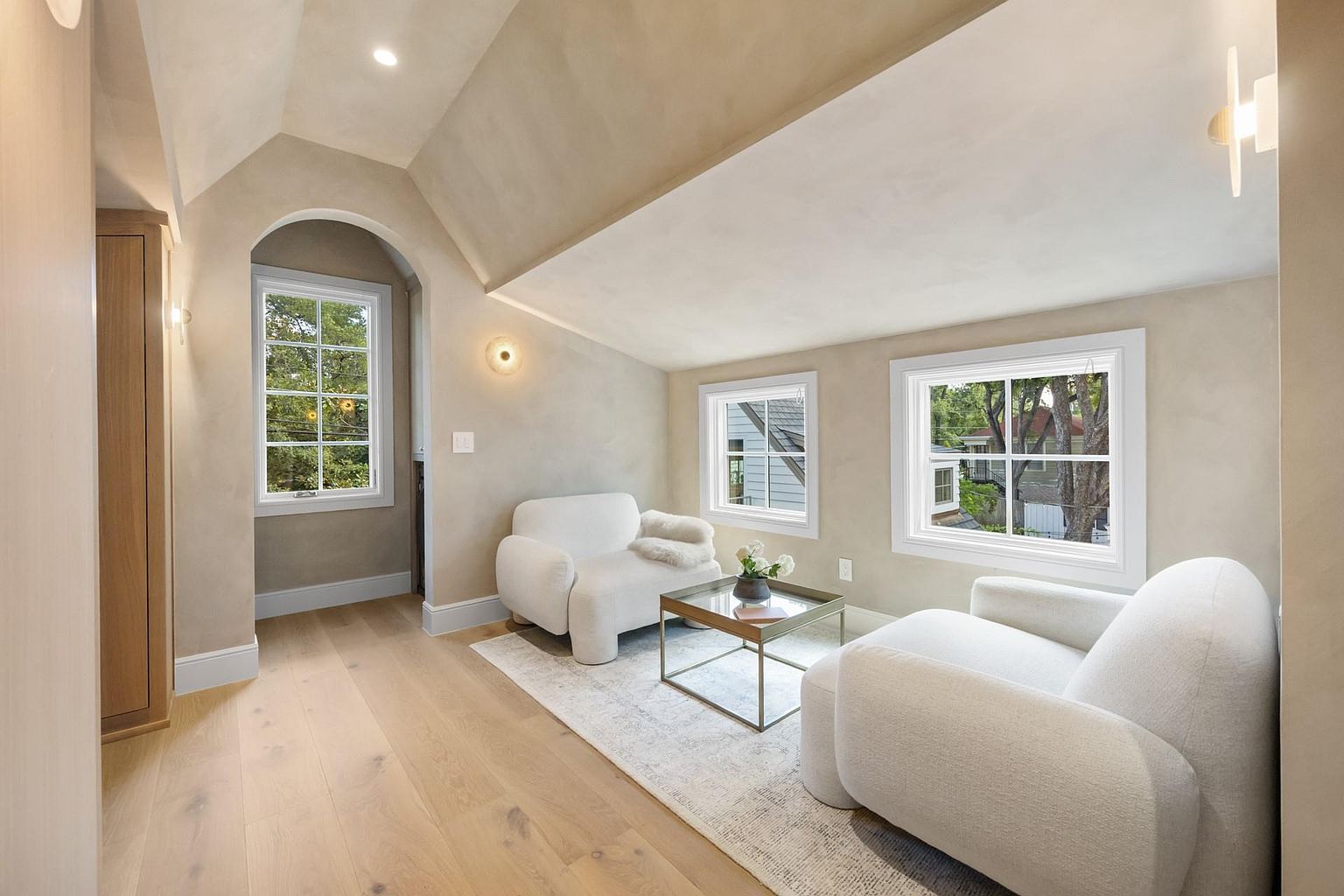 This is an interior shot of a cozy living room featuring two white armchairs, a glass-topped coffee table, and a light-colored area rug. The room has a neutral color palette with light wood flooring and textured walls, complemented by natural light streaming through the windows. The space exudes a comfortable and inviting atmosphere, perfect for relaxation.