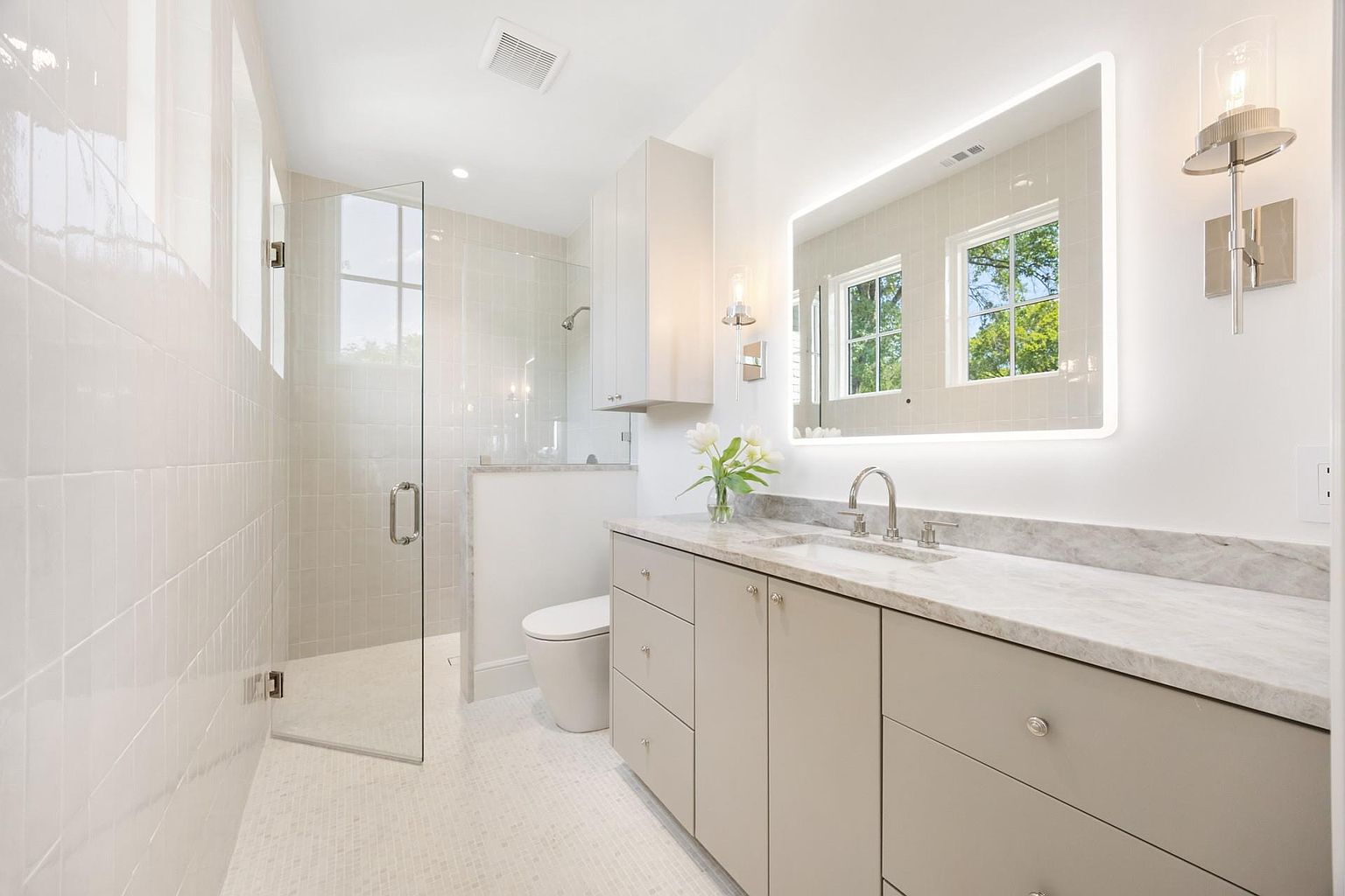 This is a bright and modern primary bathroom featuring a glass-enclosed shower, a floating vanity with a marble countertop, and a large, illuminated mirror. The neutral color palette and clean lines create a spa-like atmosphere. The floor is tiled with small white tiles.