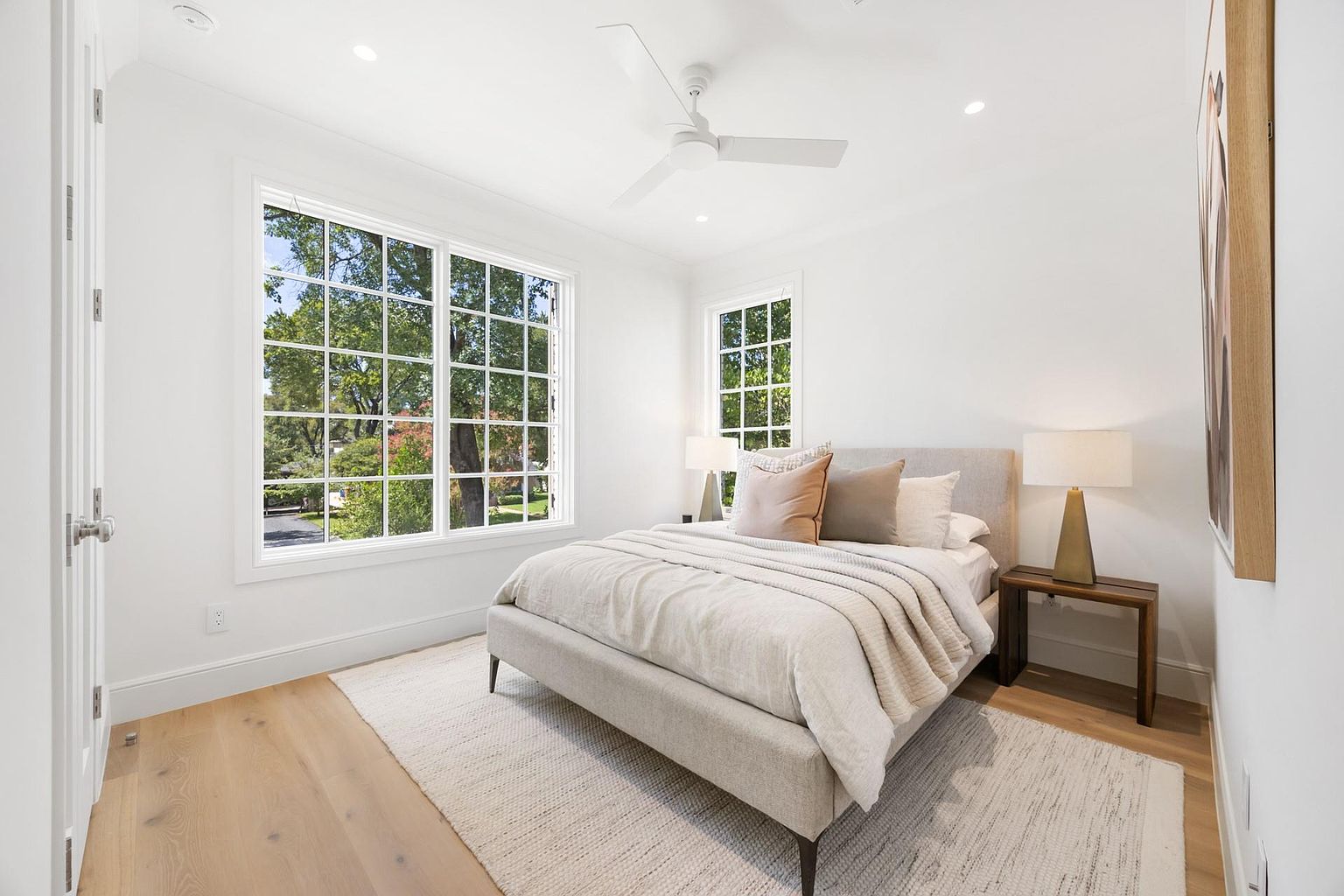 This is a bright and airy primary bedroom featuring a large window with a view of lush greenery. The room is decorated in neutral tones, with a light gray bed frame, soft bedding, and a woven rug. A wooden side table with a lamp adds a touch of warmth to the space, creating a serene and inviting atmosphere.