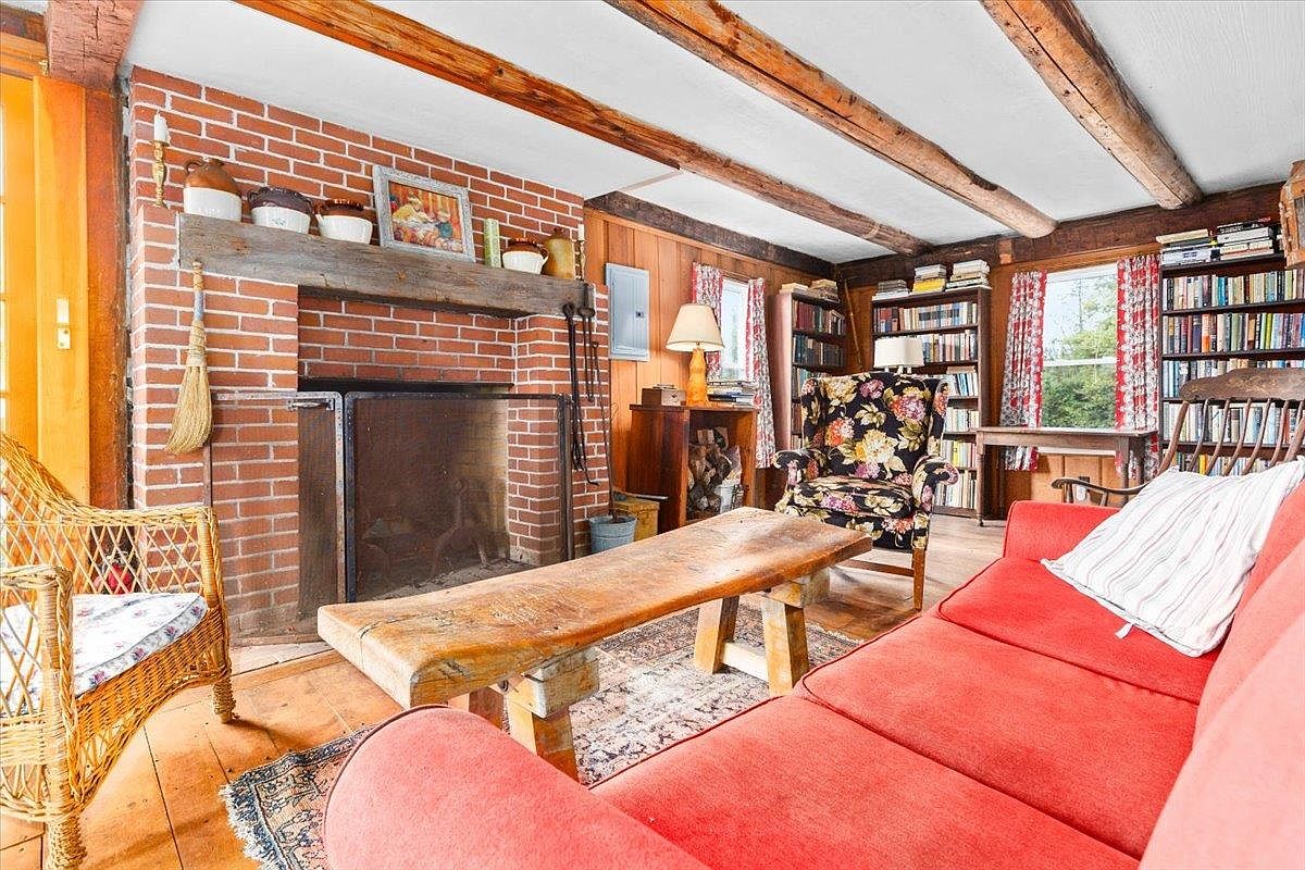 This cozy living room features a prominent brick fireplace with a rustic wood mantel and a heavy metal screen. The space is furnished with a vibrant red sofa, a floral-patterned armchair, and a natural wood coffee table, all set upon a patterned area rug. Exposed wooden ceiling beams and wood-paneled walls contribute to the room's warm, rustic aesthetic, while built-in bookshelves provide ample storage.