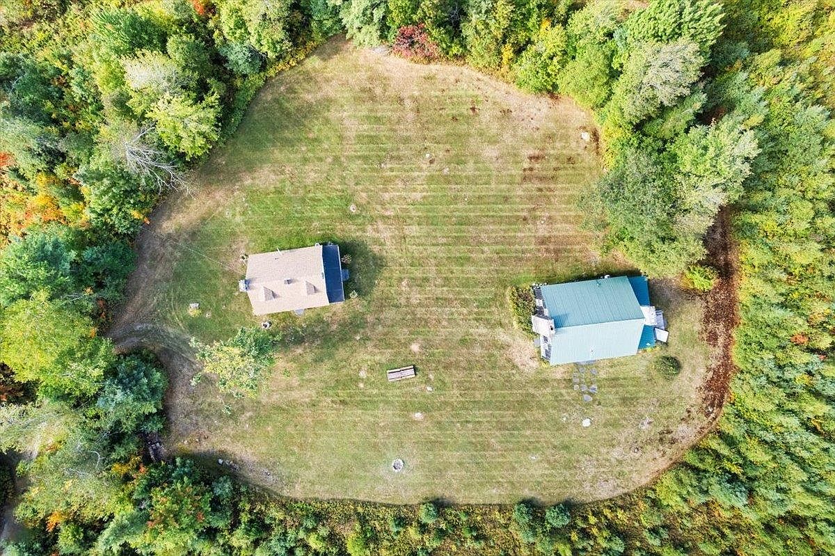 This high-angle aerial perspective captures a secluded property nestled within a dense, lush forest. The clearing features two distinct structures situated on a well-maintained lawn, with a small fire pit area positioned between them. The surrounding woodland provides a sense of privacy and natural beauty, making this an ideal setting for a quiet retreat.