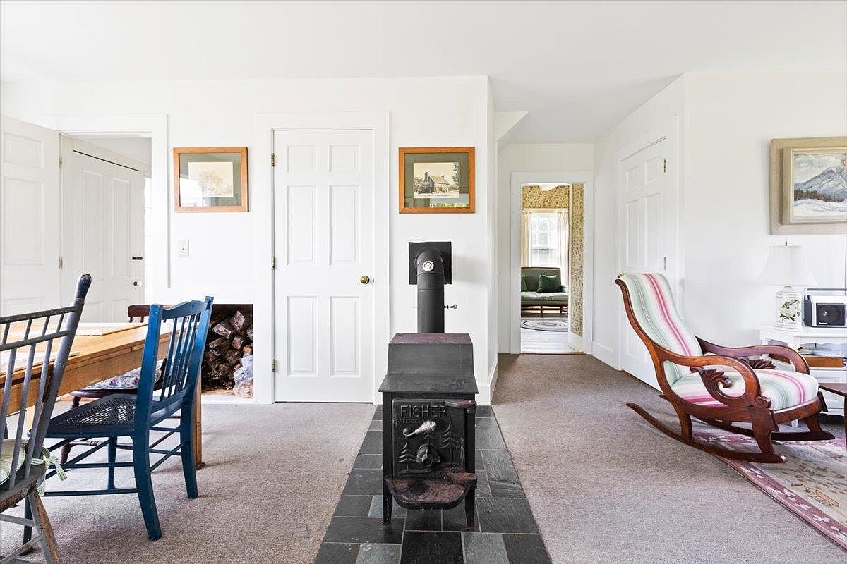 This cozy living area features a vintage Fisher wood-burning stove set on a slate hearth, serving as a central focal point. To the left, a dining space with blue chairs is visible, while a striped rocking chair sits to the right, creating a comfortable and inviting atmosphere. The room connects to an adjacent space through a doorway, showcasing a traditional home layout with neutral walls and carpeted flooring.