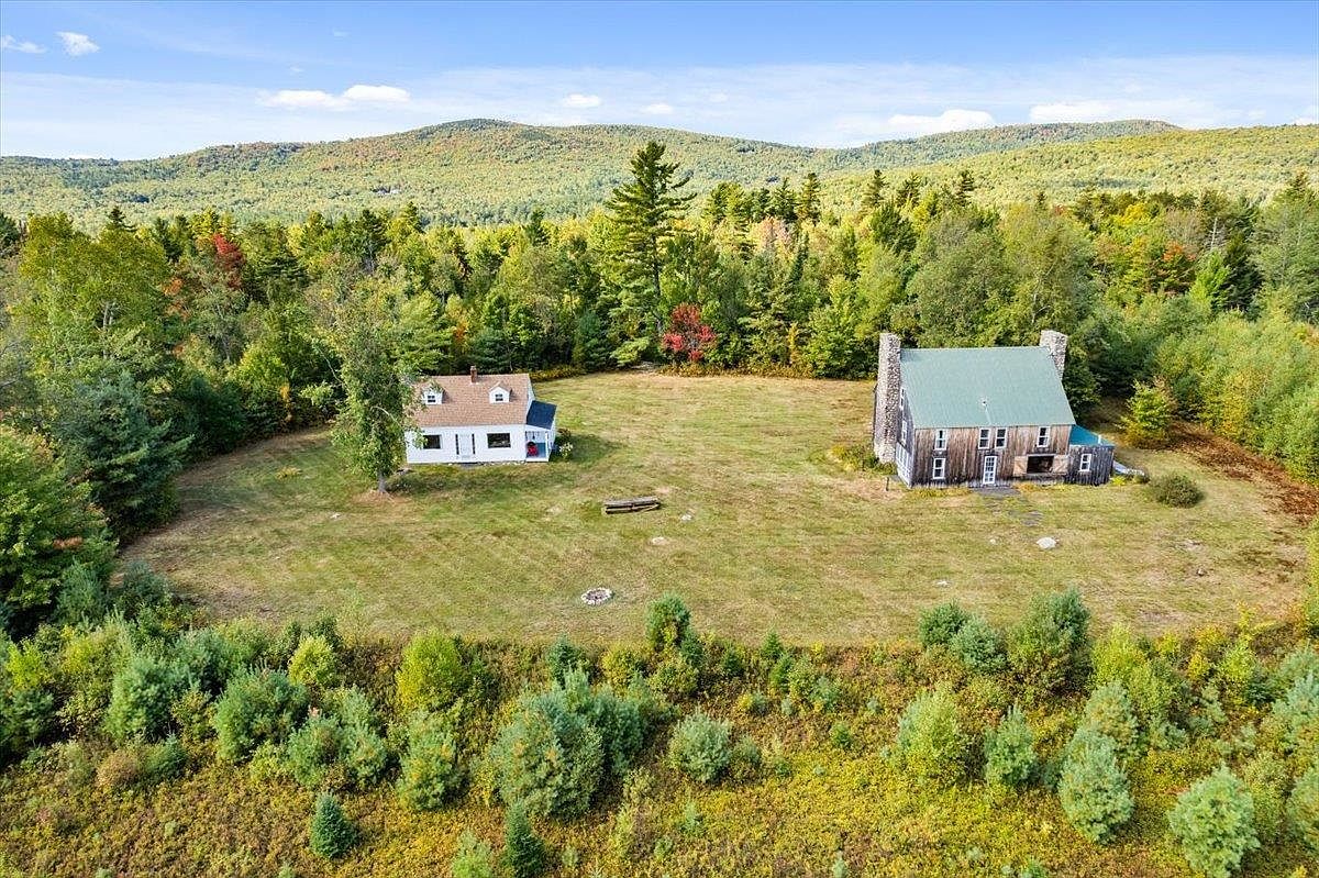 This aerial perspective captures a rural property featuring two distinct structures situated on a large, open grassy clearing surrounded by dense forest. To the left is a modest white house with a brown roof, while to the right stands a larger, rustic barn-style building with a green roof and a prominent stone chimney breast. The scene is set against a backdrop of rolling hills covered in autumn foliage, creating a serene and secluded atmosphere.