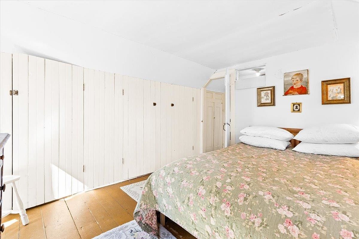This bedroom features a cozy, traditional aesthetic with light-colored wood paneling covering the walls and a sloped ceiling. A bed with a floral-patterned quilt and white pillows serves as the focal point, while framed artwork hangs on the wall above. The room is finished with warm-toned wood flooring and a small doorway leading to an adjacent space.