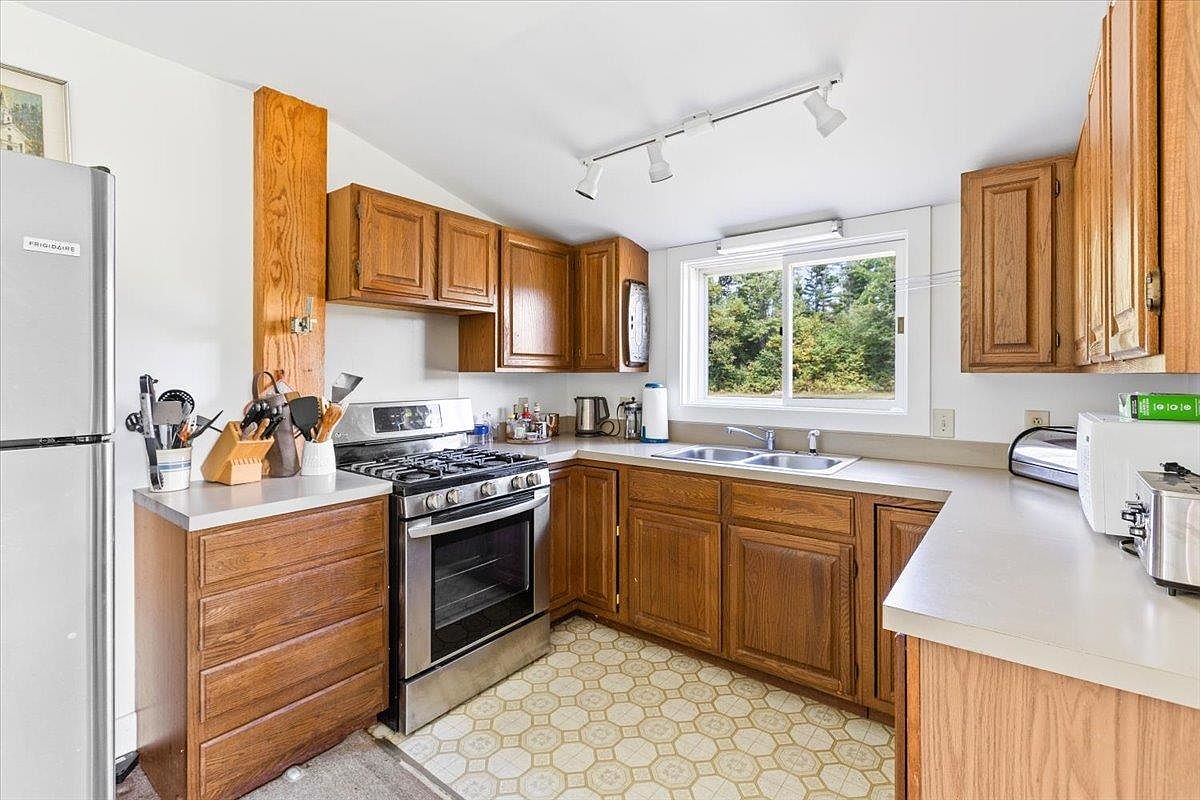 This kitchen features traditional wood cabinetry, a stainless steel gas range, and a double-basin sink positioned beneath a window that provides natural light. The space includes a light-colored countertop, a patterned tile floor, and track lighting mounted on the ceiling. A tall, vertical wood accent panel adds a rustic touch to the corner near the refrigerator.