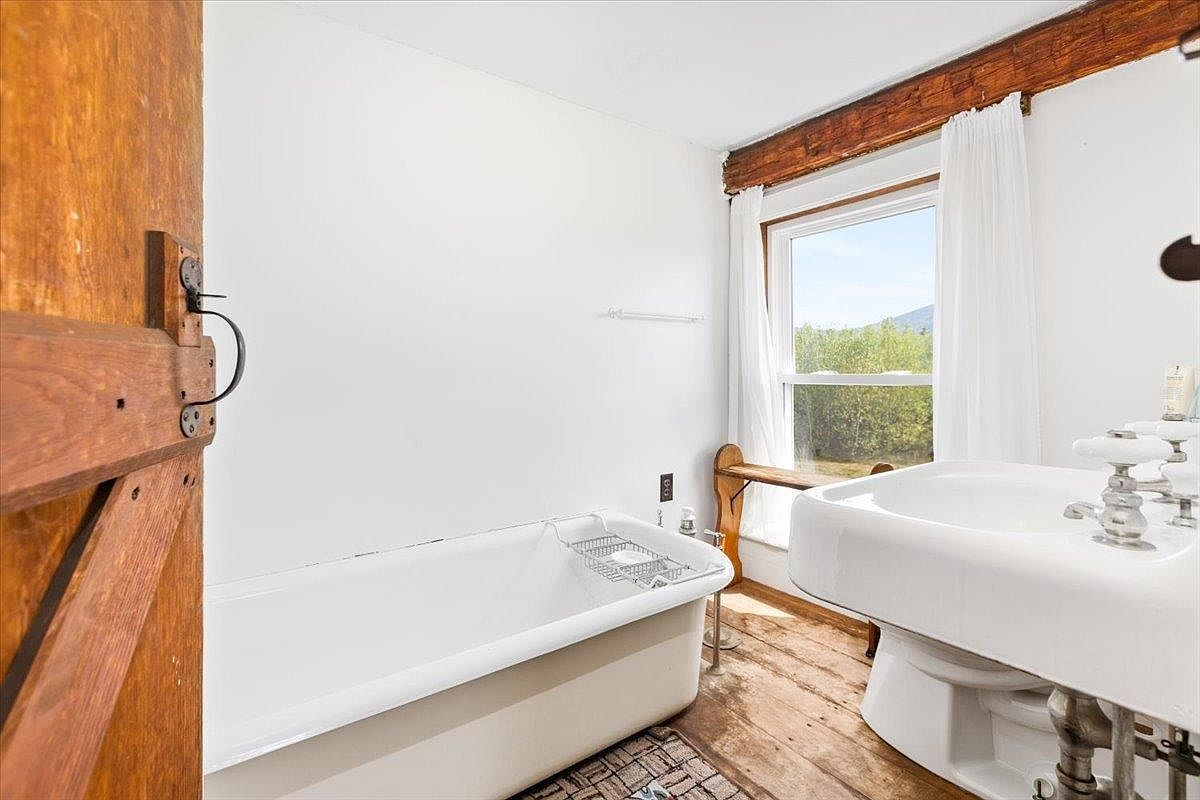 This rustic-style bathroom features a classic clawfoot tub positioned against a white wall, complemented by a pedestal sink and warm wood flooring. A large window with white curtains allows natural light to fill the space, highlighting the exposed wooden ceiling beam. The room offers a charming, vintage aesthetic with a view of the outdoors.