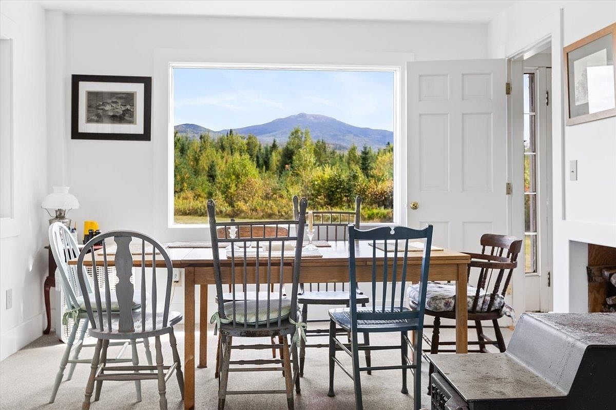 This bright dining room features a wooden table surrounded by a mix of painted and natural wood chairs, positioned to take advantage of a large picture window framing a scenic mountain view. The room is finished with white walls and neutral carpeting, creating a clean and airy atmosphere. A door to the right provides access to an adjacent space, while a portion of a wood-burning stove is visible in the foreground, adding a cozy, rustic touch to the interior.