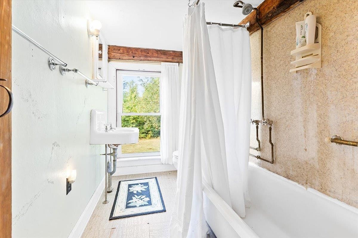 This rustic-style bathroom features a wall-mounted sink, a bathtub with a shower curtain, and exposed wooden ceiling beams. A window provides natural light and a view of the outdoors, while a patterned rug sits on the floor. The space combines vintage charm with functional elements, creating a cozy and character-filled atmosphere.