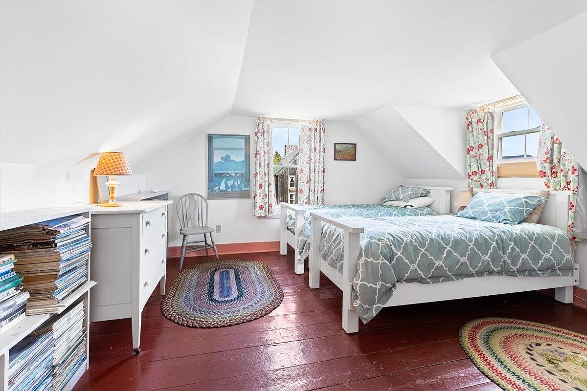 This charming attic bedroom features slanted ceilings and warm, dark-stained wood flooring, creating a cozy and inviting atmosphere. The room is furnished with two twin beds dressed in patterned blue bedding, a white dresser with a lamp, and a wooden chair, all accented by two round braided rugs. Natural light streams in through the dormer windows, which are framed by floral curtains, highlighting the bright white walls and classic cottage aesthetic.