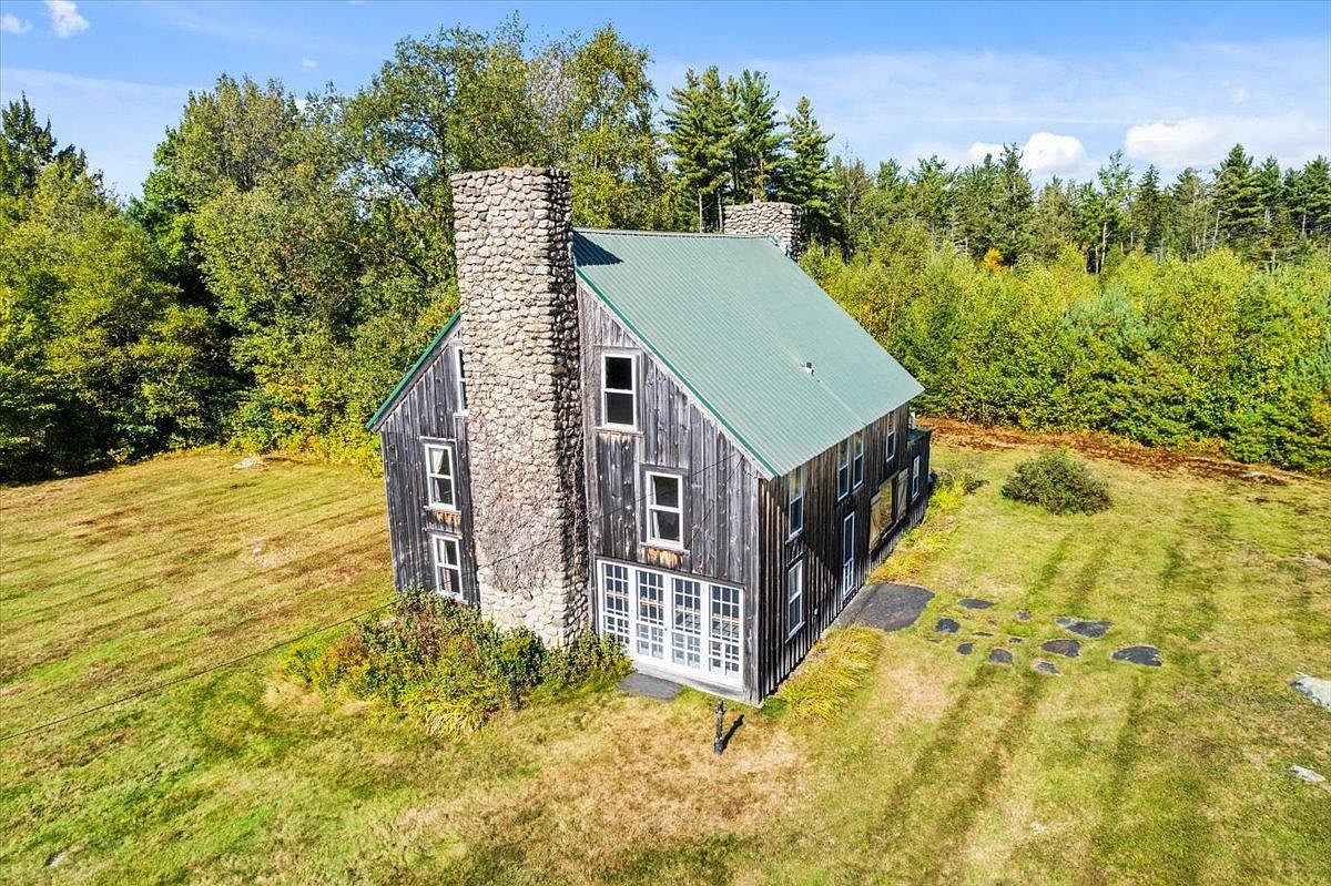 This aerial perspective captures a rustic, multi-story home featuring weathered wood siding and a prominent stone-clad chimney breast. The structure is topped with a green metal roof and is surrounded by a sprawling, open grassy lawn bordered by dense, mature trees. The composition highlights the home's secluded, natural setting and its distinctive architectural character.