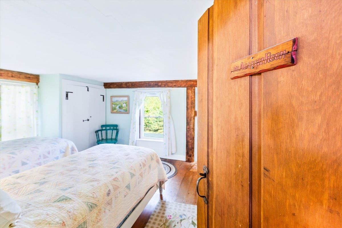 This cozy bedroom features two twin beds with light-colored patterned quilts and a rustic aesthetic defined by exposed wooden ceiling beams. A wooden door in the foreground displays a decorative sign reading 'Agassiz Room,' while the room itself is brightened by natural light from windows dressed in simple curtains. The space has a charming, cottage-like feel with light blue walls and warm wood flooring.