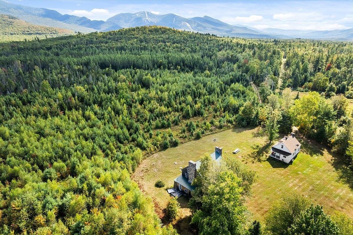 This aerial perspective captures a secluded property nestled against a vast expanse of dense, lush forest and rolling mountain ranges. The scene features two distinct structures situated on a sprawling, open grassy lot, highlighting the home's private and tranquil setting. The composition emphasizes the expansive natural landscape and the peaceful, remote character of the estate.