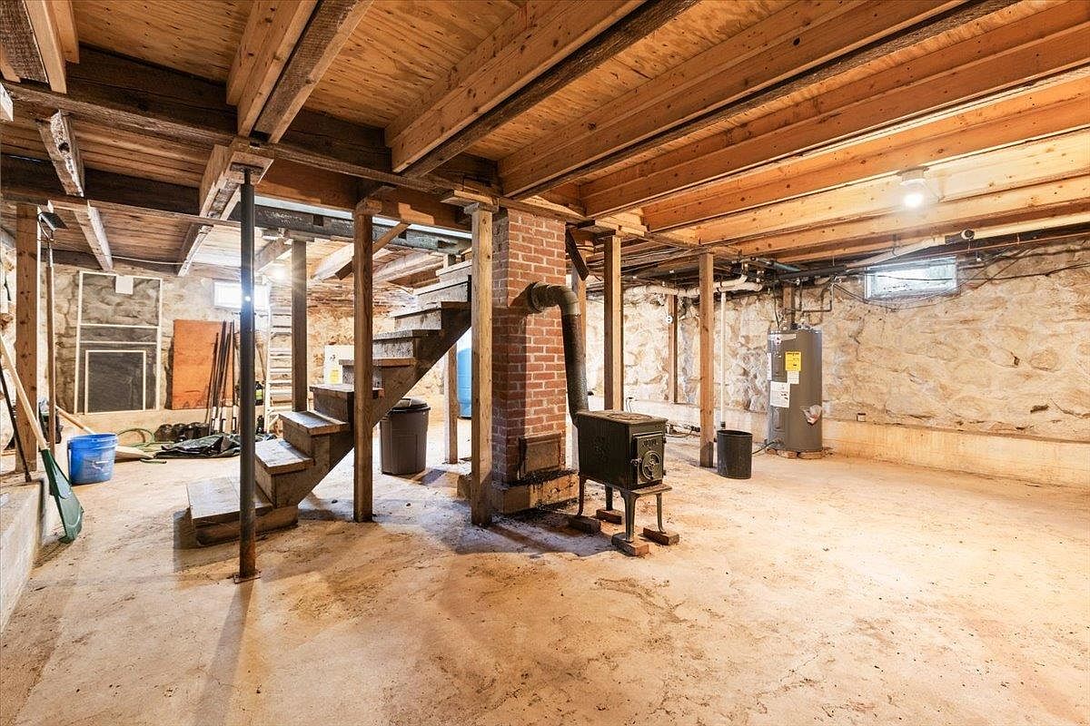 This unfinished basement features exposed wooden ceiling joists, stone foundation walls, and a concrete floor. A central brick chimney breast supports a wood-burning stove, while a wooden staircase provides access to the upper level. The space is illuminated by a single overhead light and small, high-set windows, creating a rustic, utilitarian atmosphere.