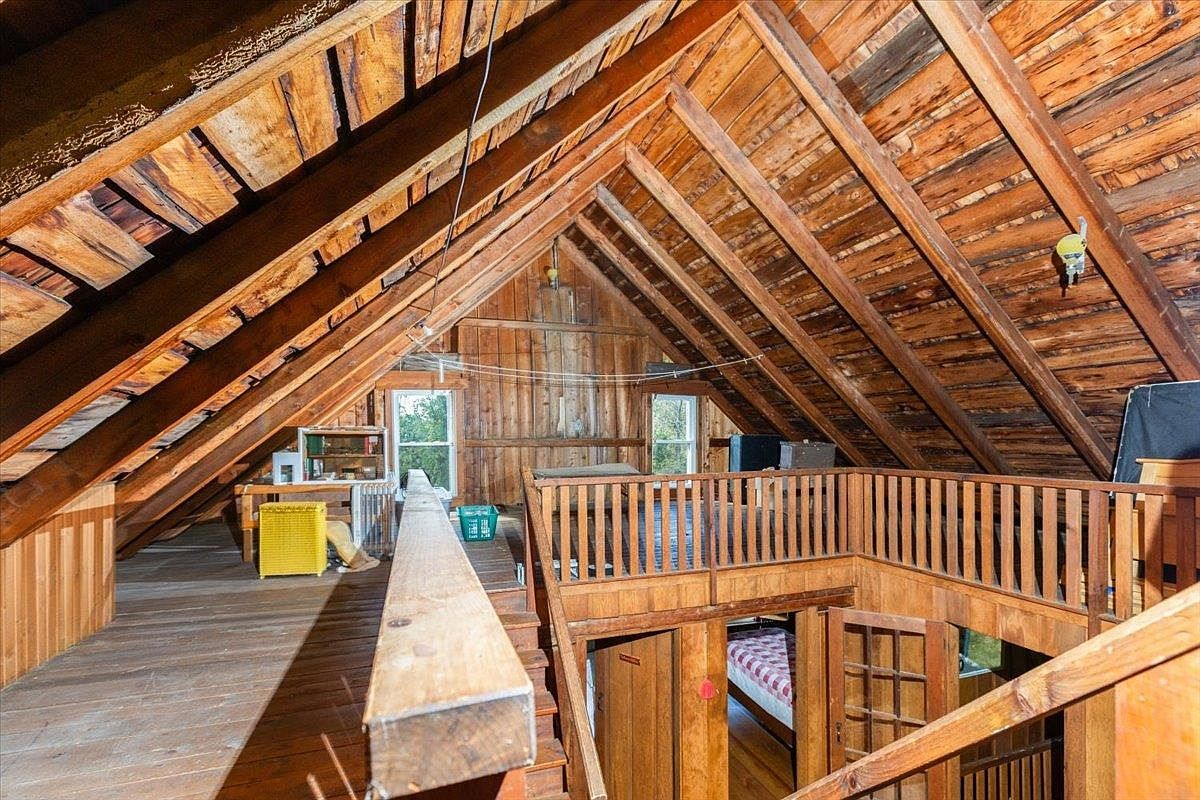 This rustic attic space features exposed wooden rafters and plank flooring, creating a warm, cabin-like atmosphere. The area includes a lofted landing with a wooden railing overlooking a lower level, two windows that provide natural light, and a prominent wooden beam extending across the foreground. The open-concept design highlights the structural timber framing and provides a versatile space with a view into the room below.