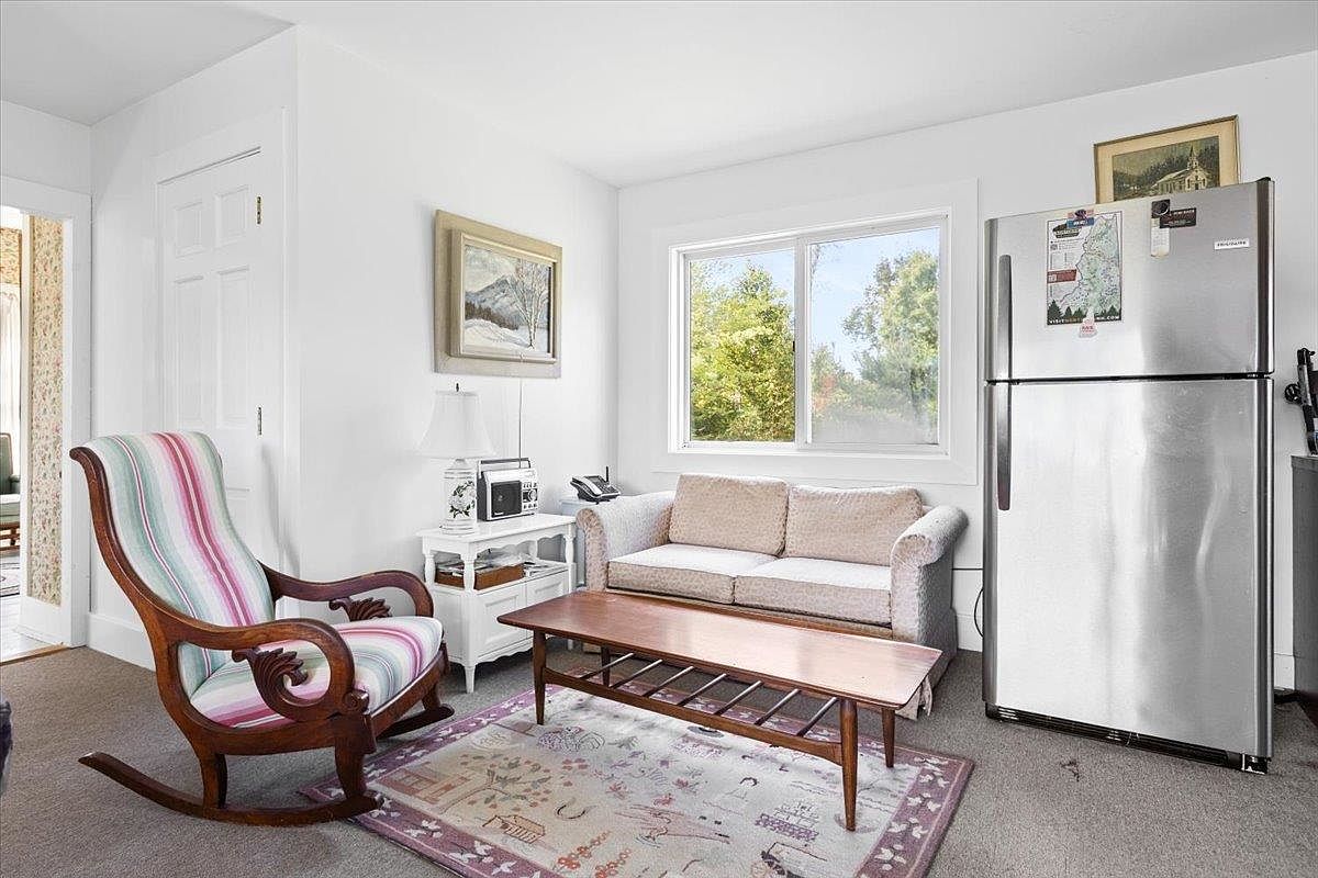 This cozy living area features a classic wooden rocking chair with striped upholstery positioned near a small side table and a light-colored loveseat. A low-profile wooden coffee table sits atop a patterned area rug, while a large stainless steel refrigerator is integrated into the corner of the room. The space is brightened by a window that allows natural light to fill the room, creating a comfortable and functional living environment.