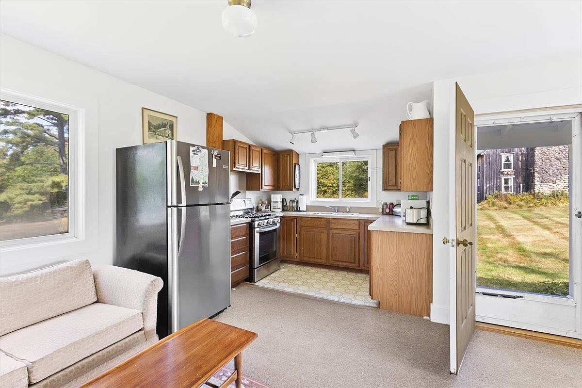 This open-concept kitchen features warm-toned wood cabinetry, stainless steel appliances, and a functional layout that flows into a small living area. A patterned tile floor defines the kitchen workspace, while the adjacent living area is carpeted and includes a neutral-toned sofa and a wooden coffee table. The space is brightened by natural light from multiple windows and an open door that provides a clear view of the exterior yard.