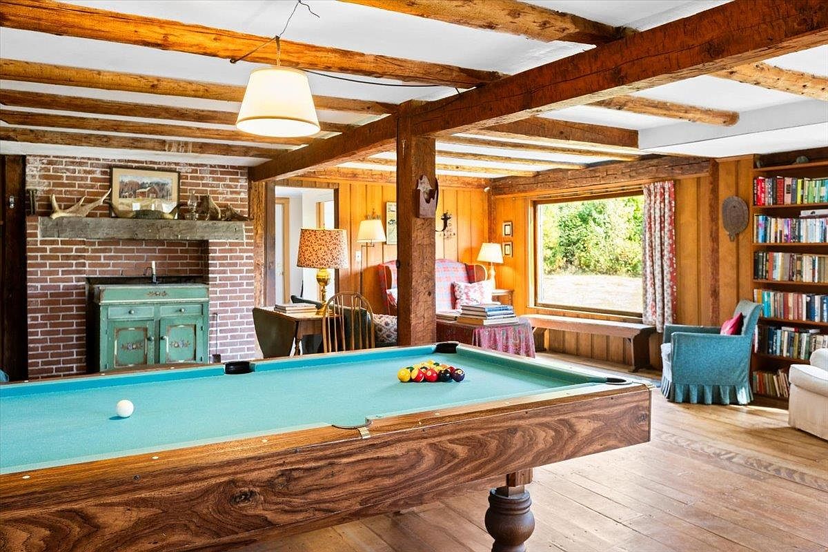 This rustic living space features a prominent pool table in the foreground, set against a backdrop of exposed timber beams and warm wood-paneled walls. A brick chimney breast with a wooden mantel serves as a focal point, while a large window provides natural light and a view of the outdoors. The room is furnished with a comfortable blue armchair and a built-in bookshelf, creating a cozy and inviting atmosphere.