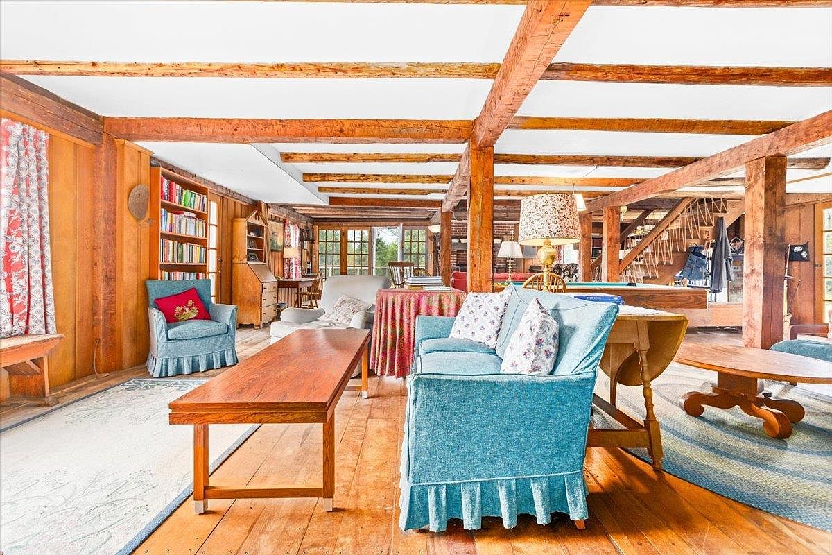 This spacious, rustic living area features exposed timber beams across the ceiling and warm wood flooring throughout. The room is furnished with a blue sofa and matching armchair, complemented by a long wooden coffee table and a bookshelf tucked into the wall. Large windows at the far end of the room allow natural light to fill the space, creating an inviting and cozy atmosphere.