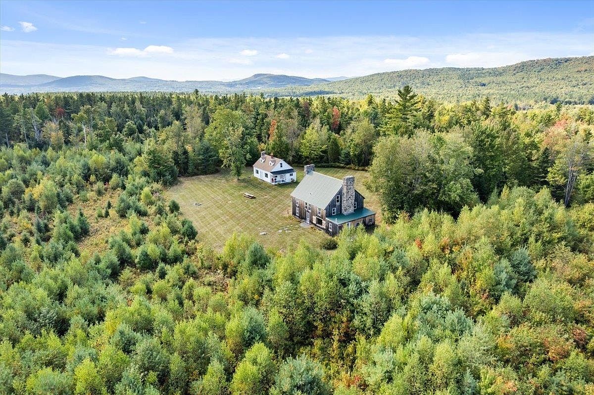 This high-angle aerial perspective captures a secluded property nestled within a dense, lush forest landscape. The scene features a large, multi-story home with a prominent stone chimney and a smaller, detached white cottage situated on a well-maintained grassy clearing. The expansive woodland setting and distant mountain range provide a serene, private atmosphere ideal for a rural retreat.