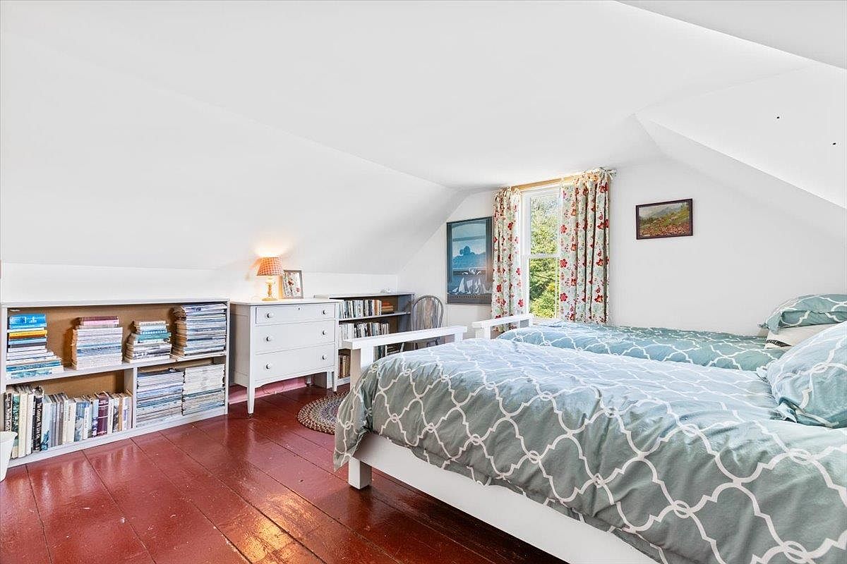 This cozy attic bedroom features a sloped ceiling and warm, dark-toned wood flooring. The room is furnished with two twin beds adorned with patterned blue and white bedding, a white dresser, and extensive built-in shelving filled with books and magazines. A window with floral curtains allows natural light to brighten the space, creating a charming and inviting atmosphere.