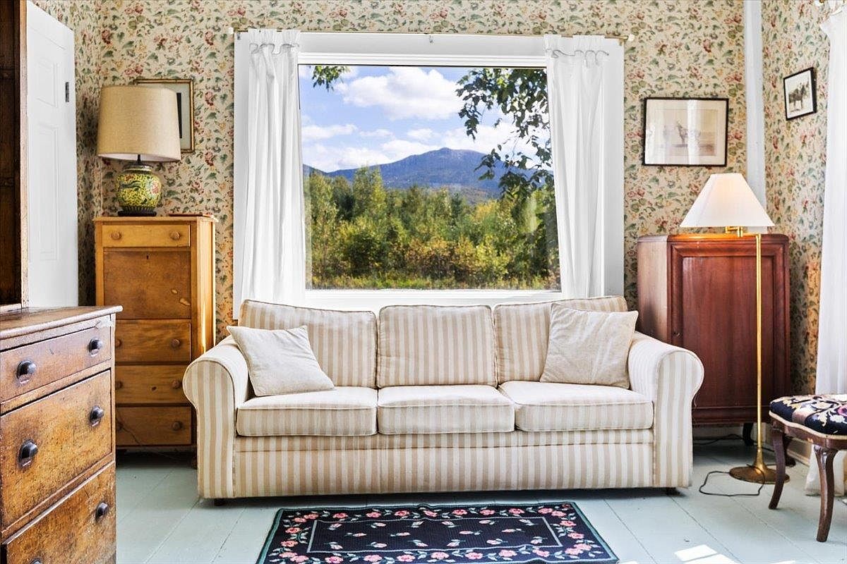 This cozy living room features a classic striped sofa centered beneath a large window that frames a scenic mountain and forest view. The space is decorated with floral wallpaper, light wood furniture, and a dark patterned area rug, creating a traditional, cottage-like atmosphere. The perspective is a straight-on, eye-level shot that highlights the inviting seating area and the natural light pouring in from the outdoors.