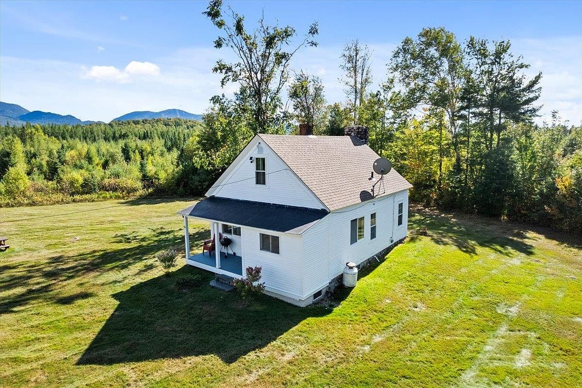 This aerial perspective captures a charming white-sided cottage nestled in a vast, grassy meadow surrounded by dense forest and distant mountains. The home features a covered front porch, a simple gable roof, and a prominent stone chimney on the rear elevation. The expansive, open landscape provides a serene and private setting, emphasizing the property's connection to the natural environment.