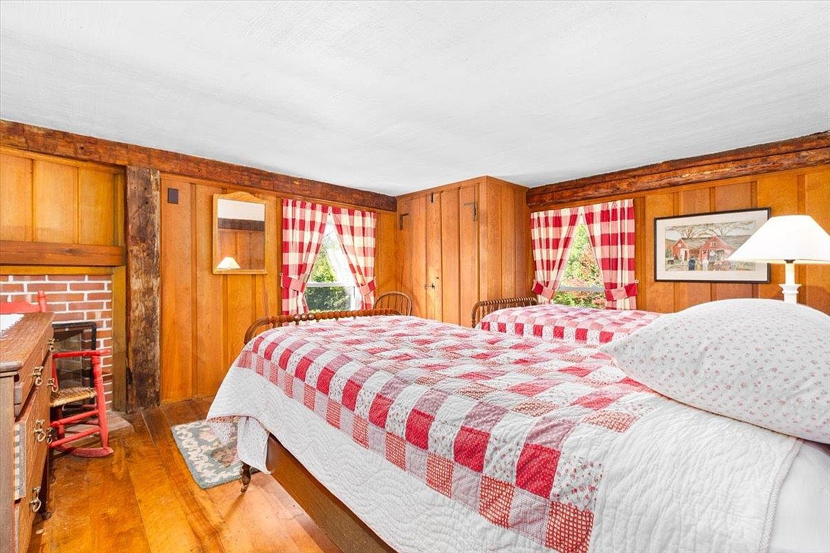 This cozy bedroom features rustic wood-paneled walls and exposed ceiling beams, creating a warm, traditional atmosphere. The room is furnished with two twin beds dressed in matching red and white checkered quilts, complemented by a brick fireplace in the corner. Large windows with matching checkered curtains allow natural light to brighten the space, highlighting the wood flooring.