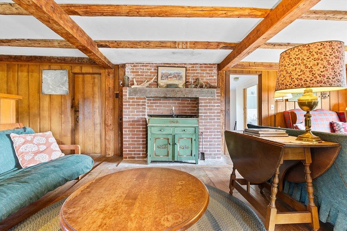 This rustic living room features exposed wooden ceiling beams and warm wood-paneled walls, creating a cozy, cabin-like atmosphere. A central brick chimney breast serves as a focal point, housing a green painted cabinet beneath a wooden mantelpiece. The space is furnished with a teal futon, a round wooden coffee table, and a drop-leaf table topped with a vintage-style floral lamp, all set upon a circular rug.
