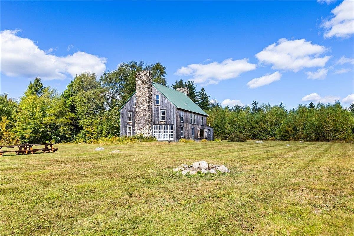 This side view captures a rustic, wood-sided home set against a backdrop of lush green trees and a bright blue sky. A prominent stone-clad chimney breast rises along the side of the structure, which features a green metal roof and multiple windows. In the foreground, a large, open grassy yard includes a picnic table and a circular stone fire pit, creating a peaceful and expansive outdoor setting.
