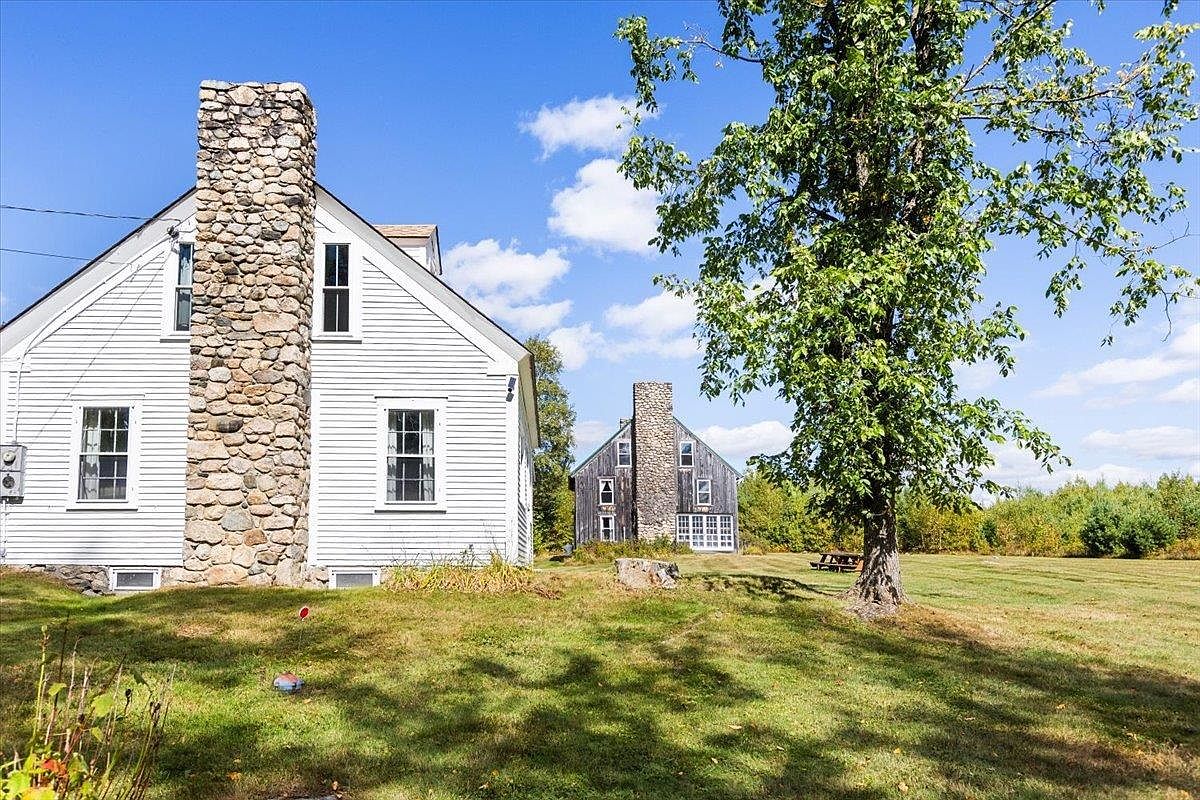 This side view captures a white-sided farmhouse featuring a prominent stone-clad chimney breast that rises along the exterior wall. A large, mature tree stands in the foreground of a sprawling, sunlit lawn, while a second, similar structure is visible in the distance. The scene conveys a peaceful, rural atmosphere with a clear blue sky and expansive green space.