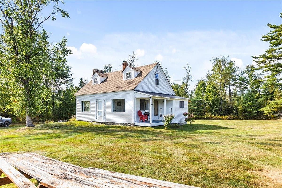 This charming white-sided cottage features a classic gabled roof with two dormer windows and a stone chimney. A small, covered front porch with a red chair provides a welcoming entry point, while the surrounding property includes a spacious, grassy lawn and mature trees. The perspective captures the home from a slightly elevated angle, highlighting its quaint, rural aesthetic and peaceful setting.