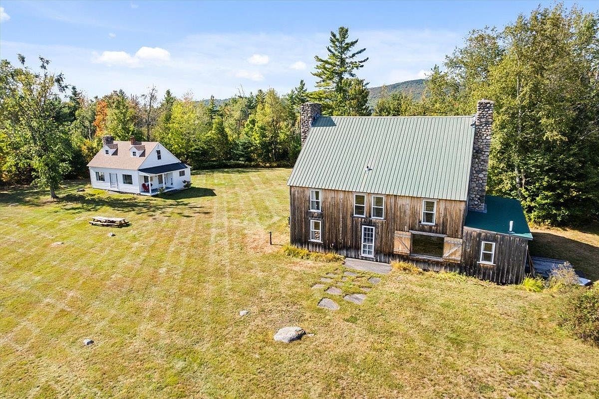 This aerial perspective captures a rustic property featuring a large, weathered wood-sided barn with a green metal roof and a stone chimney, positioned alongside a smaller, white-sided cottage. The two structures are set within a sprawling, mowed grassy field surrounded by a dense forest and rolling hills in the distance. The scene conveys a peaceful, rural atmosphere with a clear, sunny sky overhead.