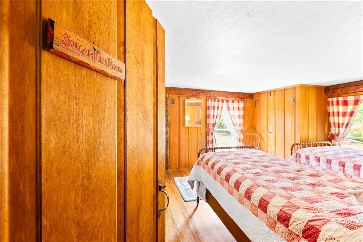 This cozy bedroom features warm wood-paneled walls and two twin beds dressed in classic red and white checkered quilts. A wooden sign labeled 'Sugar Hill Room' hangs on the door frame, adding a rustic, charming touch to the space. The room is illuminated by natural light streaming through windows adorned with matching checkered curtains, creating a quaint and inviting atmosphere.