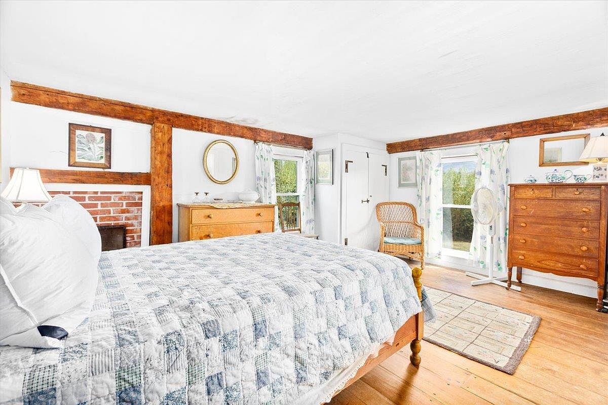 This cozy bedroom features exposed wooden ceiling beams and warm-toned wood flooring, creating a rustic and inviting atmosphere. The room is furnished with a bed dressed in a blue and white patterned quilt, a wooden dresser, and a wicker chair positioned near a window. A brick fireplace surround adds character to the space, while light-colored walls and floral curtains contribute to a bright, traditional aesthetic.