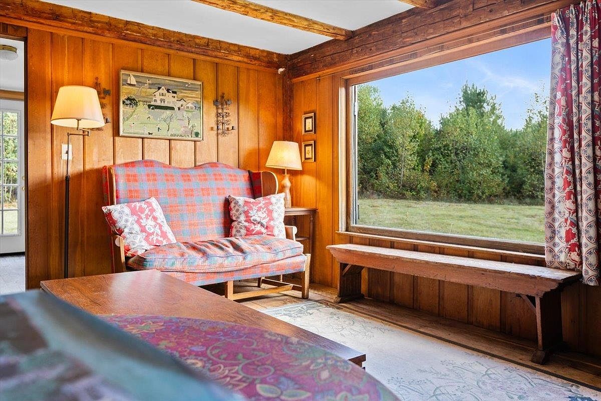 This cozy living space features warm wood-paneled walls and a rustic aesthetic, centered around a plaid-upholstered loveseat with decorative throw pillows. A large window provides a clear view of the surrounding greenery, complemented by a wooden bench placed directly beneath it. The room is accented by a floor lamp, a side table with a lamp, and framed artwork, creating a comfortable and inviting atmosphere.