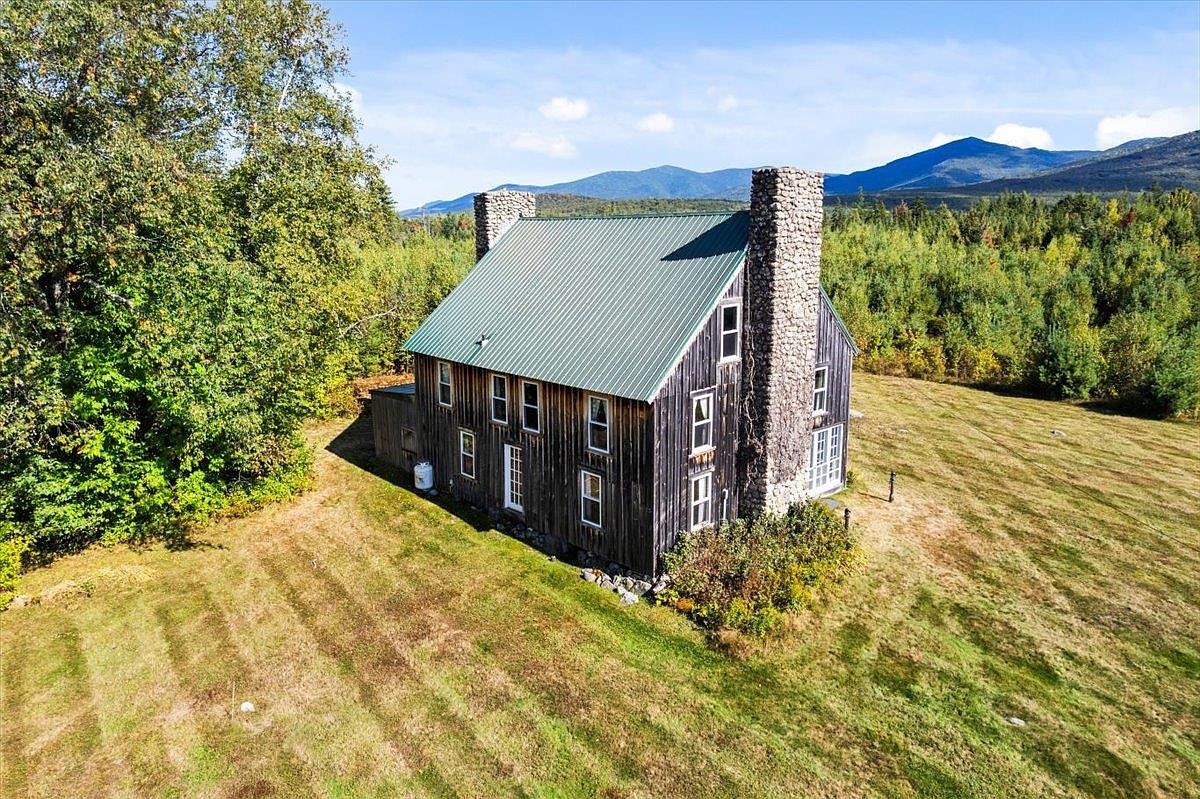 This aerial perspective captures a rustic, dark-wood-sided home set against a backdrop of rolling mountains and dense forest. The structure features a prominent green metal roof and a substantial stone-clad chimney breast on the side elevation. The property is surrounded by a vast, open grassy field, emphasizing a secluded and tranquil rural setting.