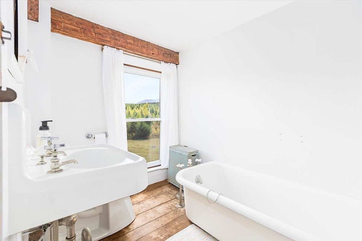 This rustic-style bathroom features a classic pedestal sink and a freestanding soaking tub set against a backdrop of white walls and warm wood flooring. A large window with white curtains allows natural light to fill the space, highlighting the exposed wooden ceiling beam. The room offers a clean, minimalist aesthetic with a view of the surrounding landscape.