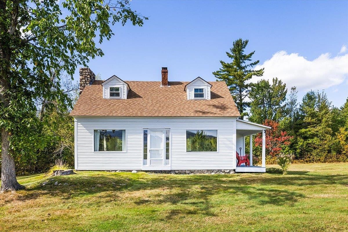 This charming white-sided cottage features a classic symmetrical facade with two dormer windows and a central front door. A stone-clad chimney rises from the side of the house, and a small covered porch on the right side provides a cozy outdoor seating area. The home is set on a grassy lawn surrounded by mature trees, creating a peaceful and inviting rural atmosphere.