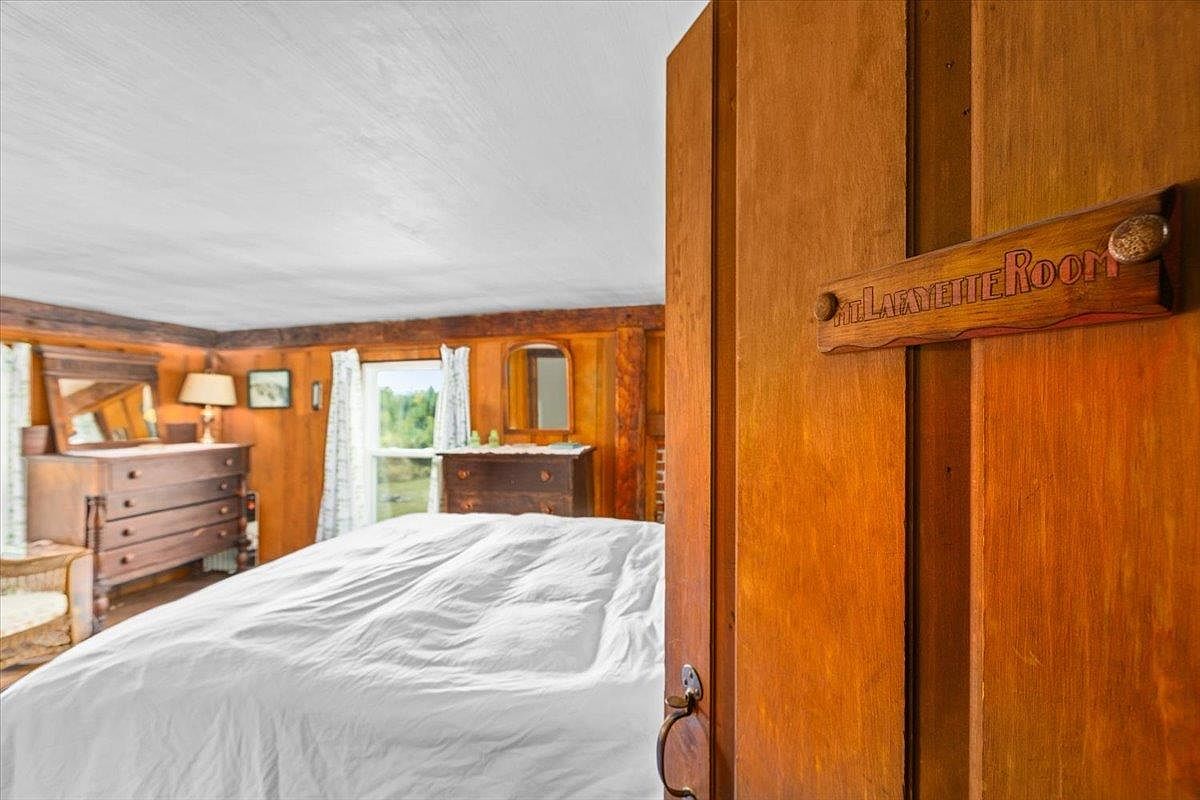 This cozy bedroom features warm wood-paneled walls and a rustic aesthetic, highlighted by a wooden sign on the door frame that reads 'Mt. Lafayette Room.' The room is furnished with a bed covered in white linens and a traditional wooden dresser topped with a mirror and lamp. The perspective is from the doorway, looking into the room, creating an inviting and intimate atmosphere.