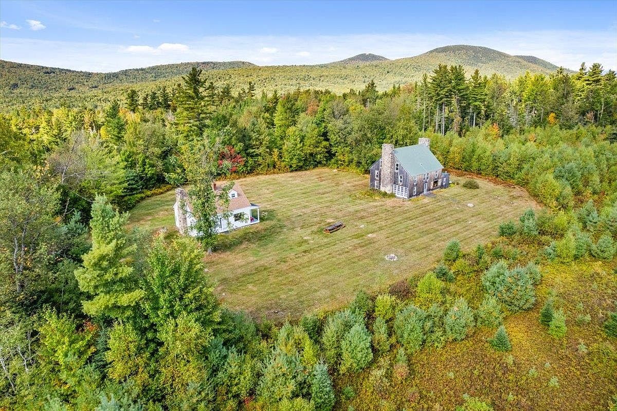 This aerial perspective captures a secluded property nestled within a dense, lush forest, featuring two distinct structures set upon a large, open grassy clearing. The larger building on the right showcases a traditional design with a prominent stone chimney, while the smaller white structure on the left offers a charming, cottage-like aesthetic. The surrounding landscape is characterized by rolling hills and a vibrant canopy of trees, creating a serene and private atmosphere.