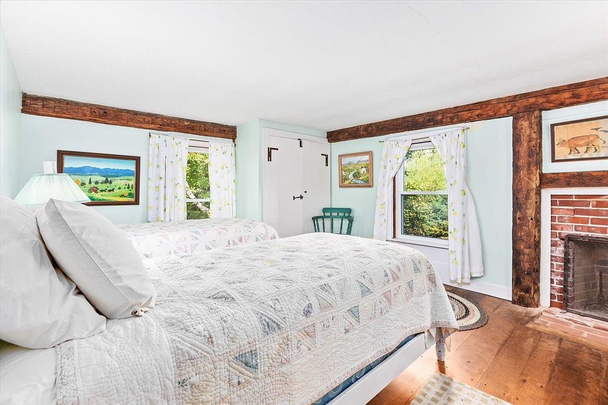 This cozy bedroom features two beds with light-colored patterned quilts and exposed wooden ceiling beams that add a rustic, historic charm. A brick fireplace with a simple mantel sits on the right, while two windows with floral curtains provide natural light to the space. The room is decorated with framed artwork and light-toned walls, creating a warm and inviting atmosphere.
