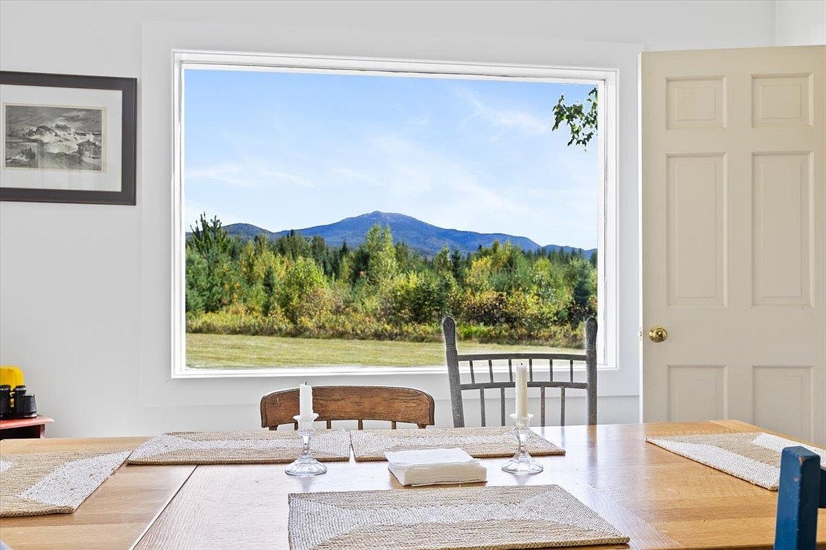 This dining room features a large wooden table set with woven placemats and simple candle holders, centered in front of a expansive window that frames a picturesque mountain and forest view. The room has a bright, airy atmosphere with white walls and a classic wooden door to the right, creating a serene and inviting space. The perspective is eye-level, focusing on the dining area as a peaceful vantage point for enjoying the natural scenery.