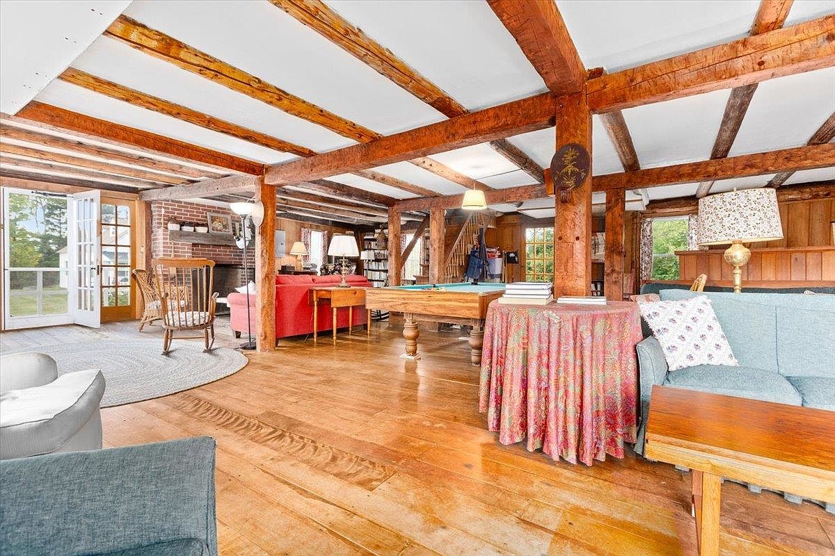 This spacious, open-concept living area features rustic exposed timber beams across the ceiling and warm wood flooring throughout. The room is furnished with a red sofa near a brick fireplace, a pool table in the center, and a cozy seating area with a blue sofa in the foreground. Large glass doors provide natural light and a view of the outdoors, creating a welcoming and expansive atmosphere.