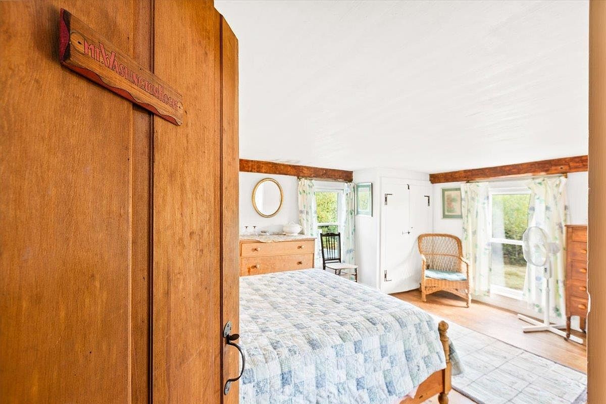 This bedroom is viewed from the doorway, revealing a cozy space with a light-colored quilt on the bed and a wooden dresser. The room features exposed wooden ceiling beams, a wicker chair near the window, and a light-toned wood floor. The perspective emphasizes the transition from the wooden door frame into the bright, airy bedroom.